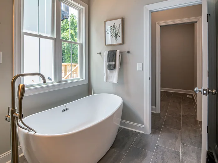 White freestanding tub by a window, gray tile floor, door to hall